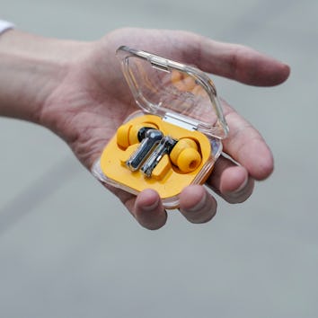 A hand holds a small transparent case with yellow wireless earbuds inside, against a blurred backgro...