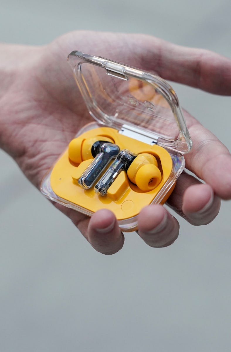 A hand holds a small transparent case with yellow wireless earbuds inside, against a blurred backgro...