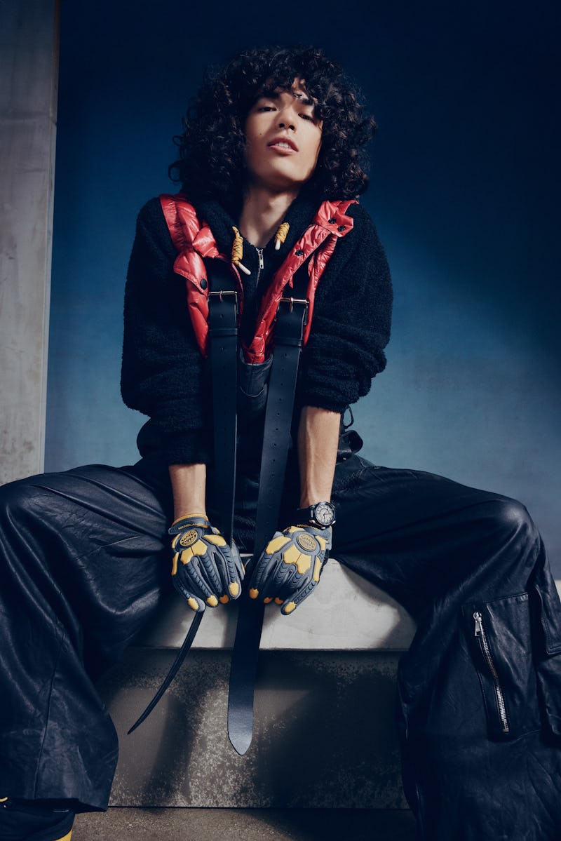 Stylish person with curly hair seated, wearing a black jacket, red vest, gloves, and leather pants, ...