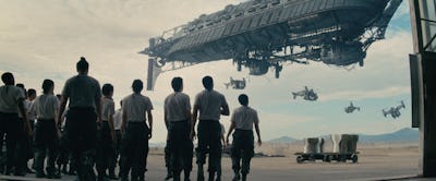 A group of people in uniform watching a spaceship as smaller craft fly by in a desert-like setting.