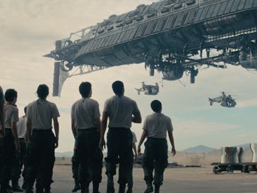A group of people in uniform watching a spaceship as smaller craft fly by in a desert-like setting.