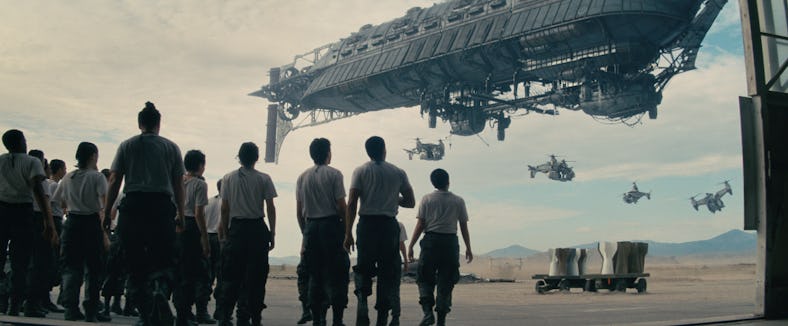 A group of people in uniform watching a spaceship as smaller craft fly by in a desert-like setting.