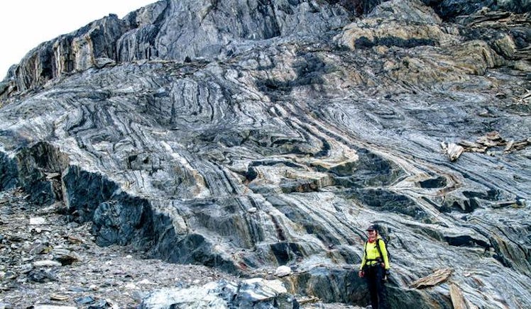 A scientist dressed as a hiker poses against a huge formation of rocks with alternating bands of dar...