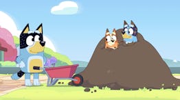 Bandit stands next to a wheelbarrow, facing Bingo and Bluey on a pile of dirt in a sunny landscape.
