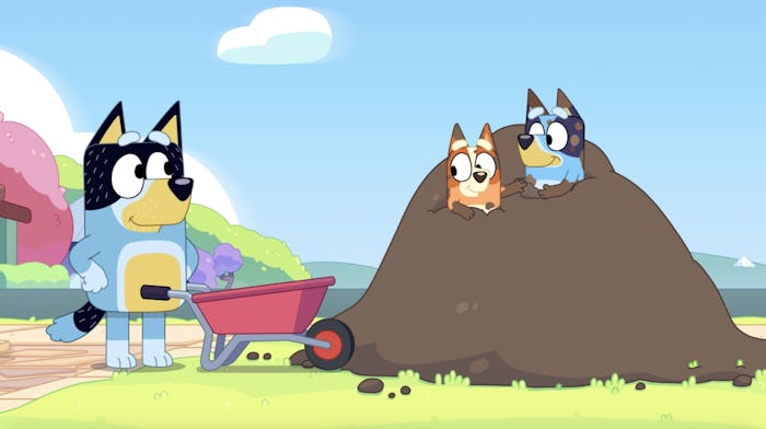 Bandit stands next to a wheelbarrow, facing Bingo and Bluey on a pile of dirt in a sunny landscape.