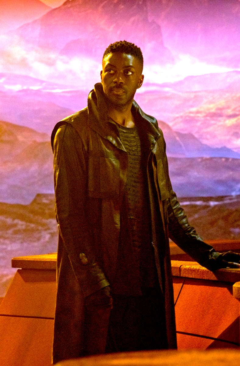 A man stands in front of a vivid alien landscape with purple skies and rocky terrain, leaning on a f...