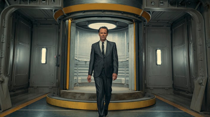 A man in a suit stands confidently in a futuristic circular doorway inside a metallic corridor.