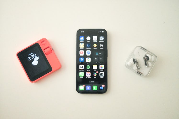 The Rabbit R1 (left) next to an iPhone 15 Pro (center) and Nothing Ear 2 wireless earbuds (right)