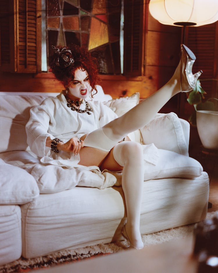 In full costume makeup, Chappell Roan sits on a couch in a white bathrobe with one leg in the air pu...
