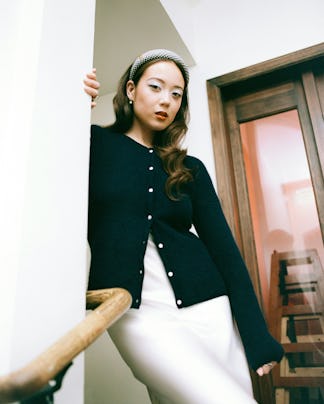 A woman in a stylish outfit poses on wooden stairs, wearing a black cardigan, white skirt, and headb...