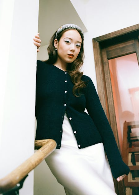 A woman in a stylish outfit poses on wooden stairs, wearing a black cardigan, white skirt, and headb...
