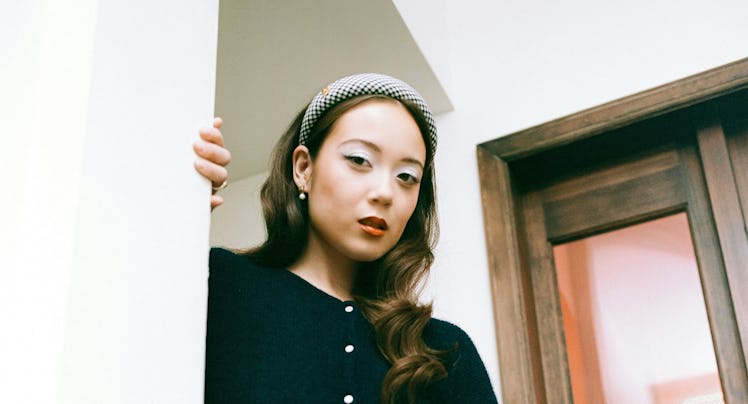 A woman in a stylish outfit poses on wooden stairs, wearing a black cardigan, white skirt, and headb...