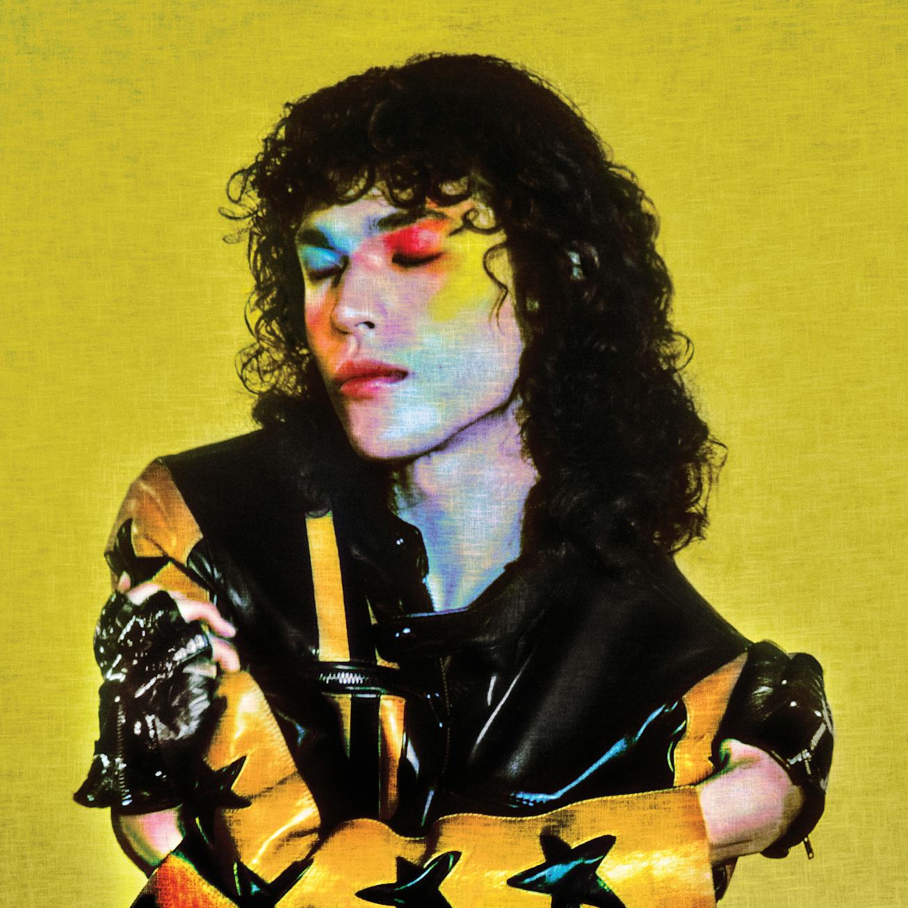 A person with curly hair and colorful makeup sits against a yellow background, wearing a shiny, patt...
