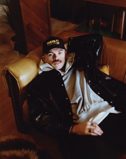 Man with a mustache wearing a cap sits relaxed in a leather chair, holding a black shiny garment.