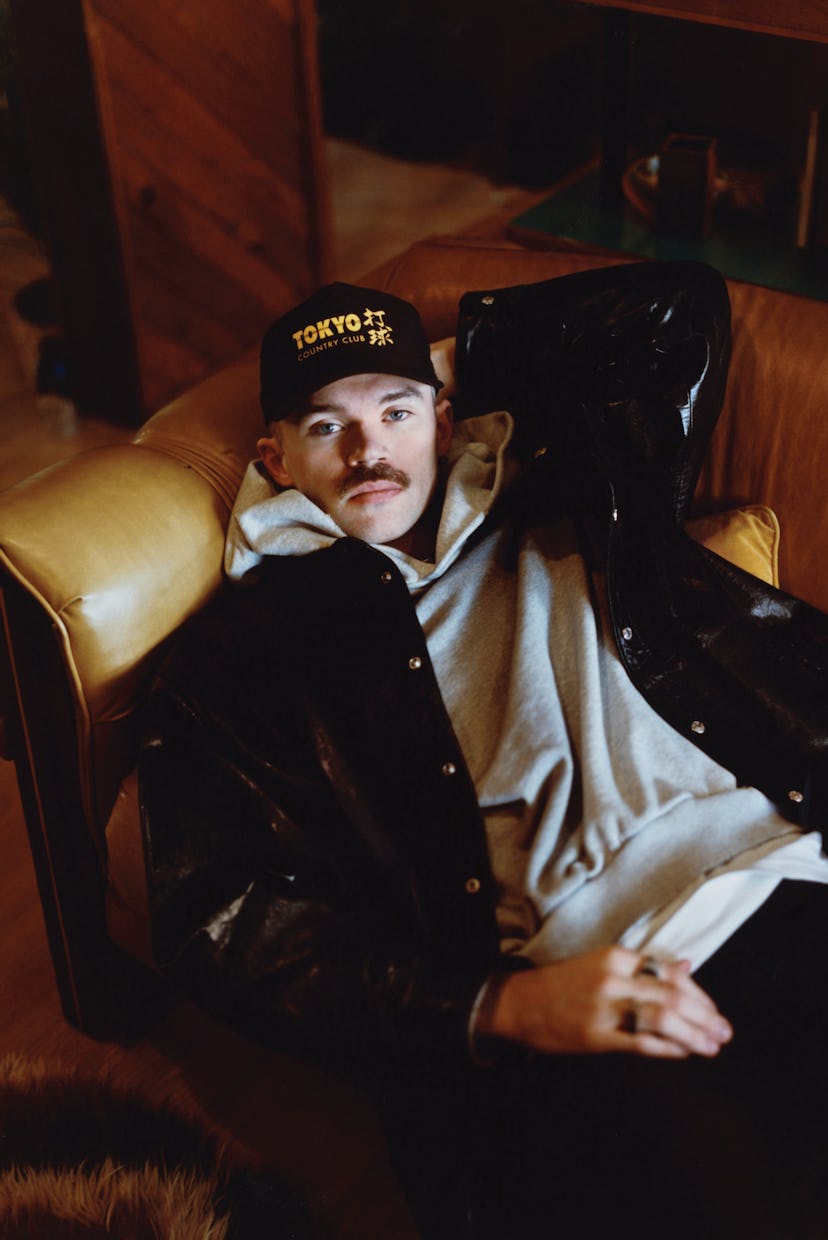 Man with a mustache wearing a cap sits relaxed in a leather chair, holding a black shiny garment.
