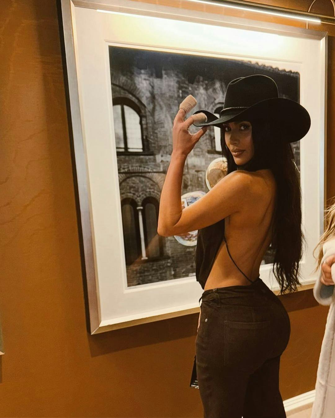 Kim Kardashian wears a plunging cowl top, pants, and cowboy hat.