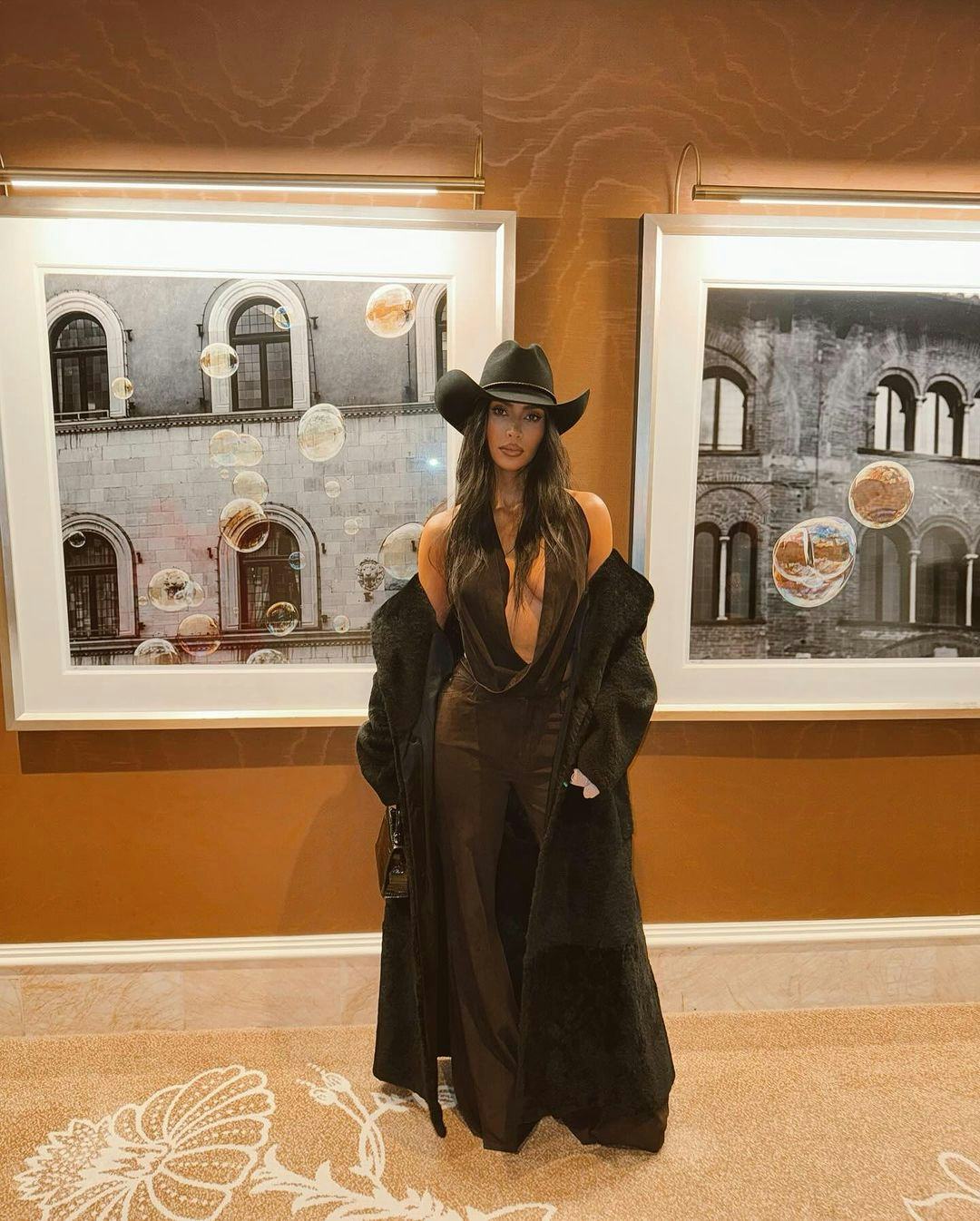 Kim Kardashian wears a plunging cowl top, pants, and cowboy hat.