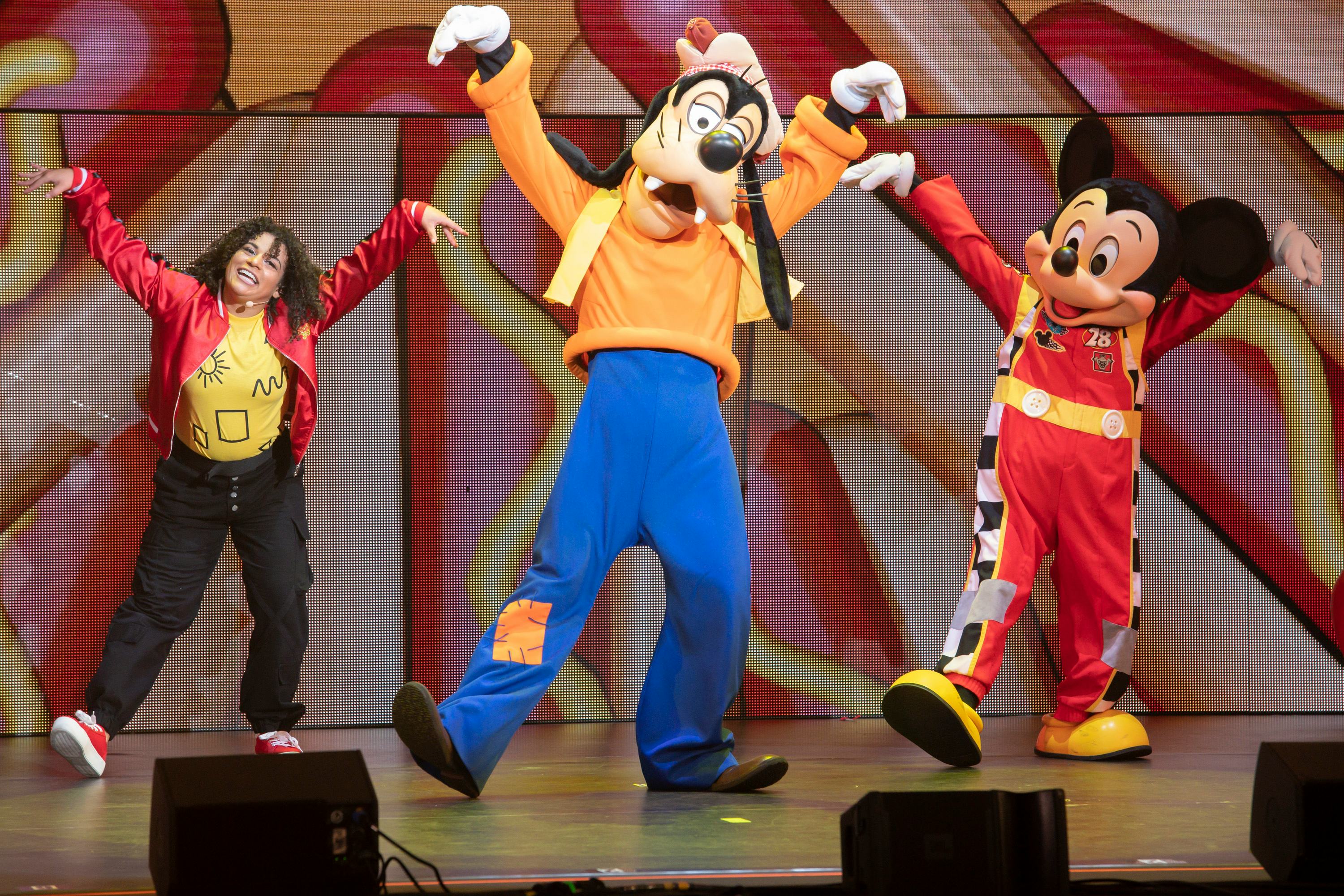 Mickey Mouse and Goofy Dance alongside a woman in a red jacket and curly hair in Disney Jr. Live.
