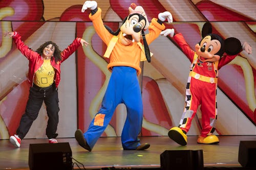 Mickey Mouse and Goofy Dance alongside a woman in a red jacket and curly hair in Disney Jr. Live.