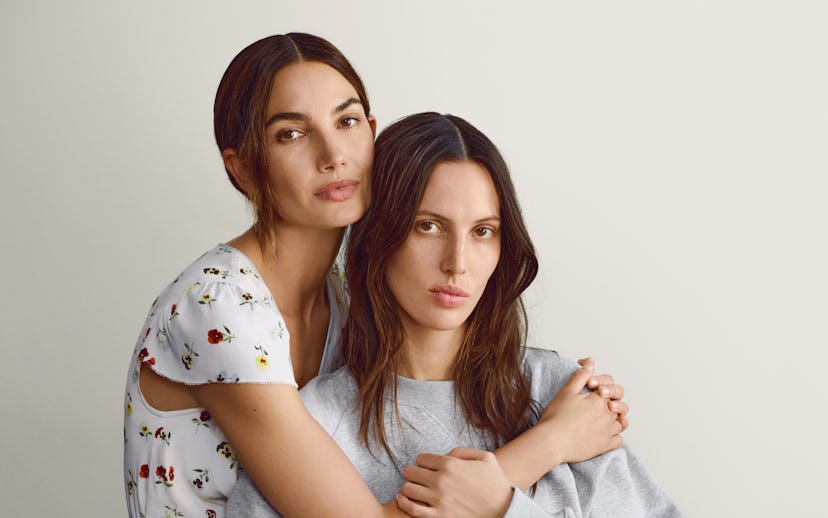 Two women embracing on a stool, one in a floral dress and the other in a gray sweatshirt, against a ...