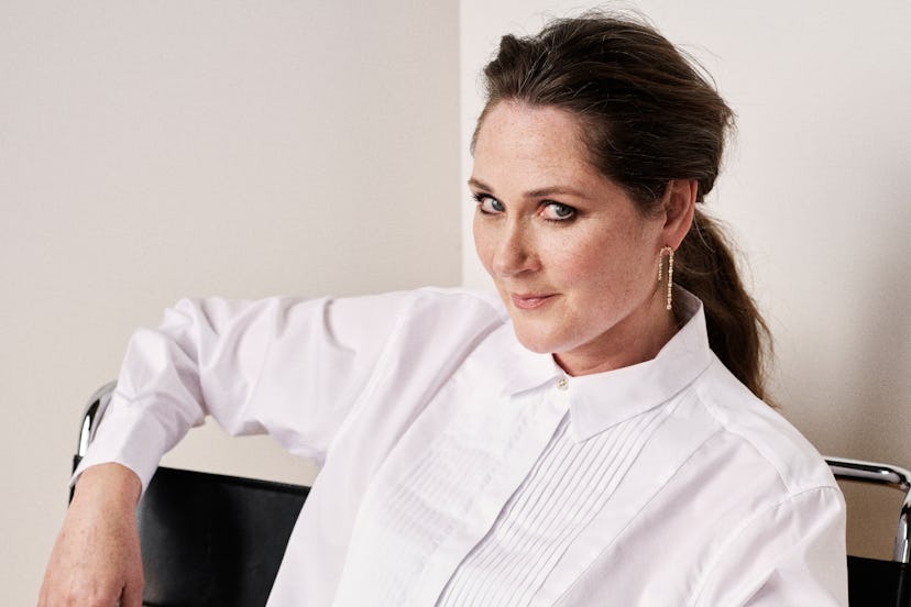 Confident woman in a white shirt sitting on a chair, looking at the camera with a subtle smile.