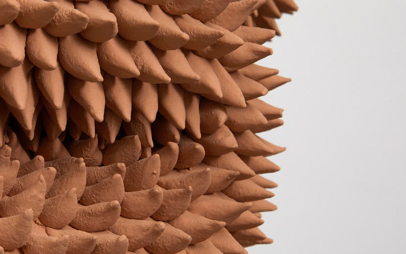 Close-up of a textured sculpture with numerous layered, terracotta colored pointy shapes against a l...