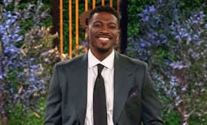 A man in a dark suit and tie smiling confidently in front of a lush, green plant background onstage.
