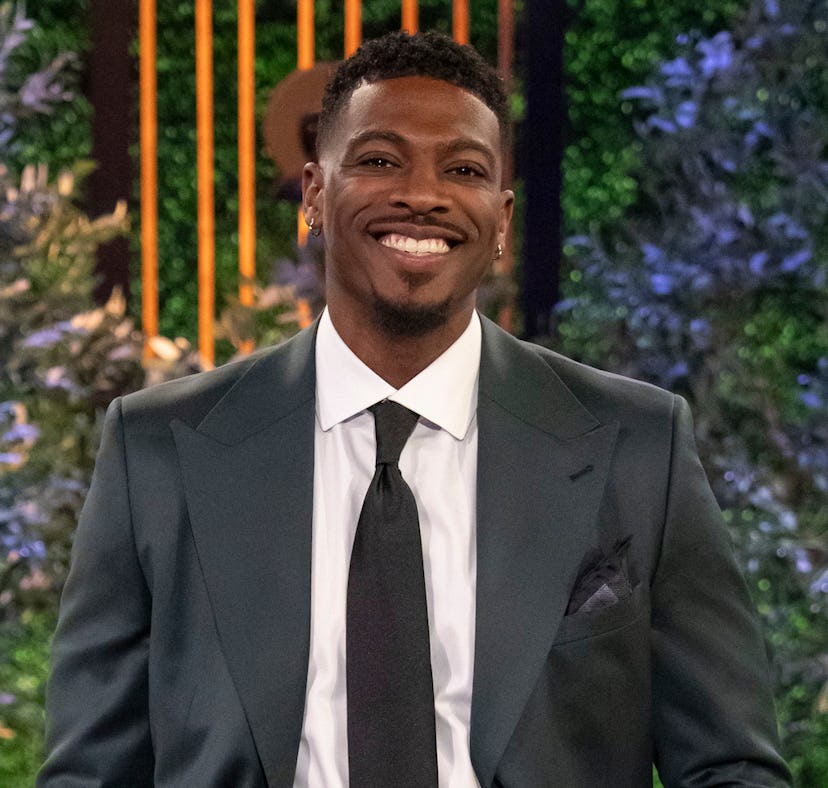 A man in a dark suit and tie smiling confidently in front of a lush, green plant background onstage.