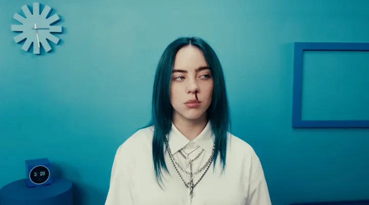 Woman with blue hair and a white shirt standing in a blue room, looking slightly to the side with a ...