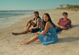 Three young adults sitting on a sandy beach looking towards the ocean with a house in the background…