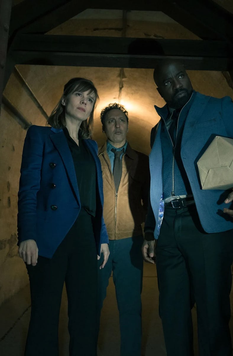 Three people, two women and a man, stand cautiously inside a dimly-lit, narrow doorway, looking inte...