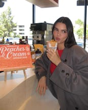 A woman in a gray blazer blows a kiss while holding a smoothie in a cafe with a promotional sign bes...