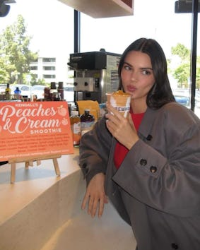 A woman in a gray blazer blows a kiss while holding a smoothie in a cafe with a promotional sign bes...