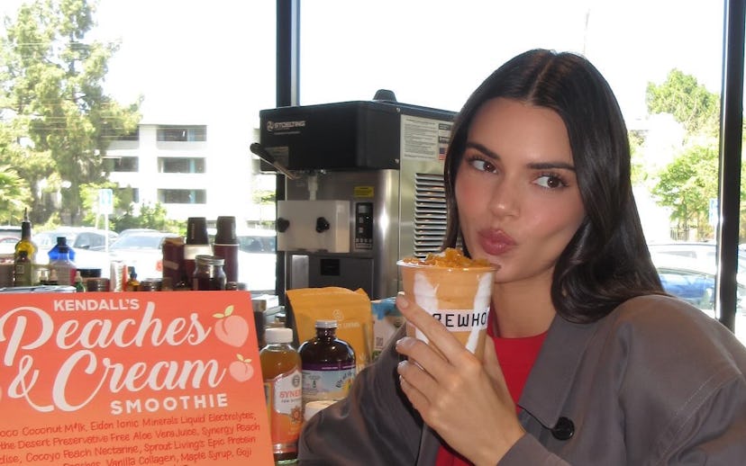 Woman in a gray blazer sitting at a cafe counter, holding a smoothie and pouting playfully towards t...