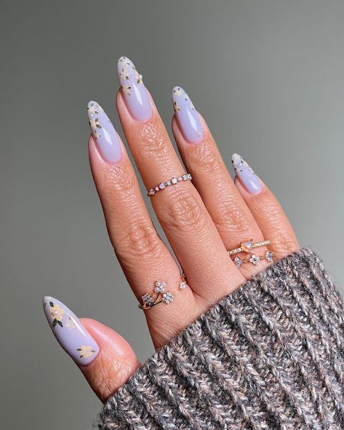 Hand showcasing long lavender nails with floral designs, adorned with rhinestone details, against a ...