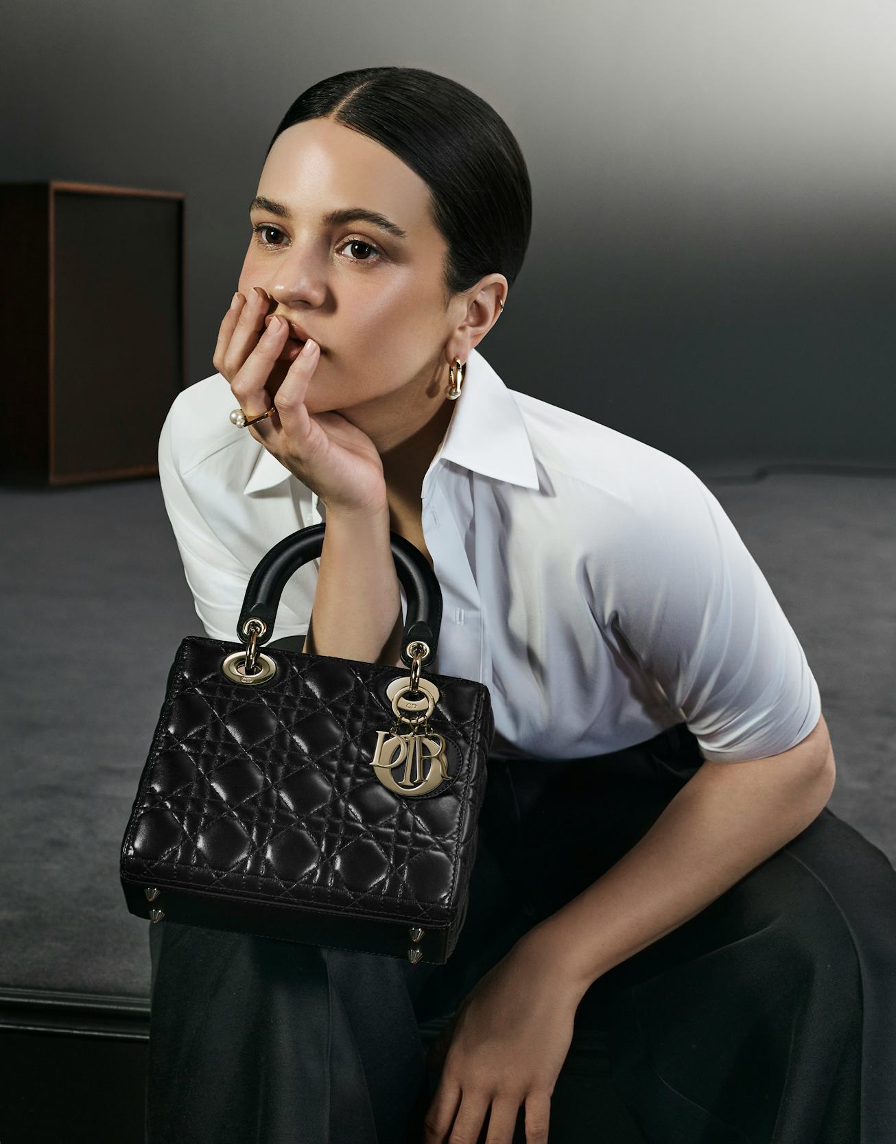 Woman in a white blouse leans forward, holding a stylish black quilted Dior handbag, with a thoughtf...