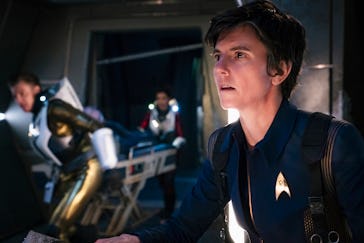A person in a blue Starfleet uniform looks intently to the side in a dim, futuristic spacecraft sett...