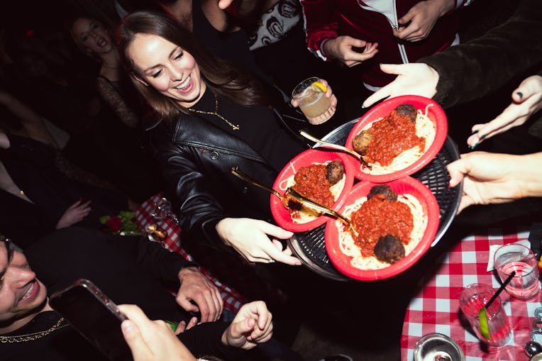 People enjoying a party, sharing plates of spaghetti and meatballs, with one person smiling and hold...