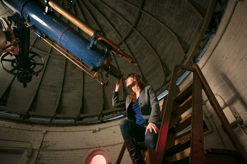A woman sits on stairs beside a large telescope inside an observatory, gazing upward thoughtfully.