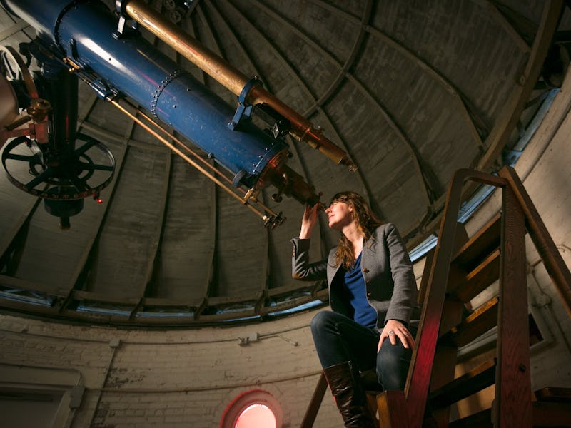 A woman sits on stairs beside a large telescope inside an observatory, gazing upward thoughtfully.