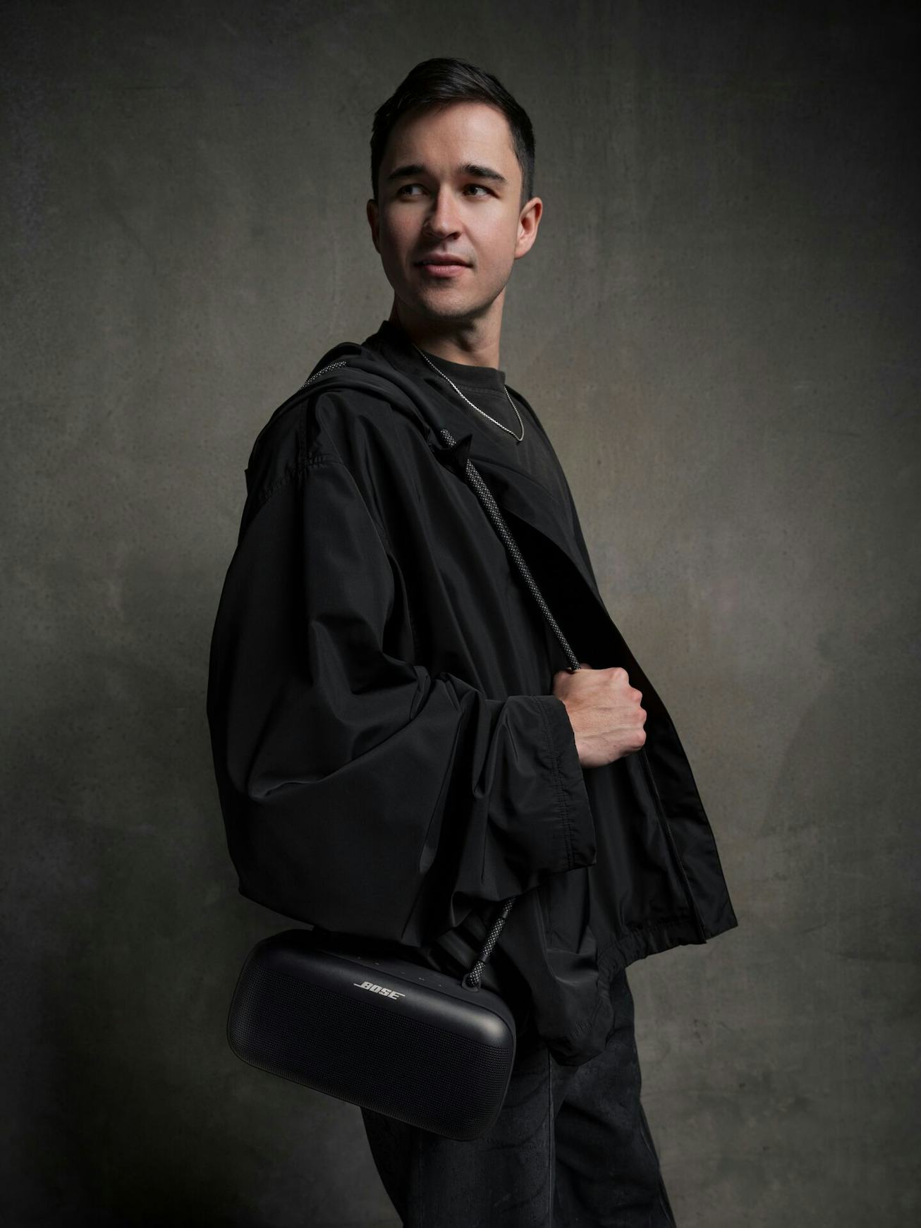 A young man in a black jacket and shirt holds a modern bag, posing against a textured grey backdrop.