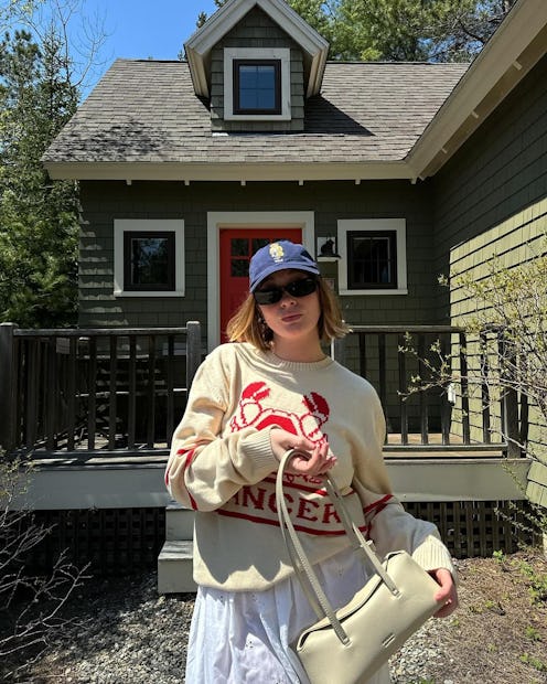 Woman in a cream sweater and cap stands in front of a green house with red door.