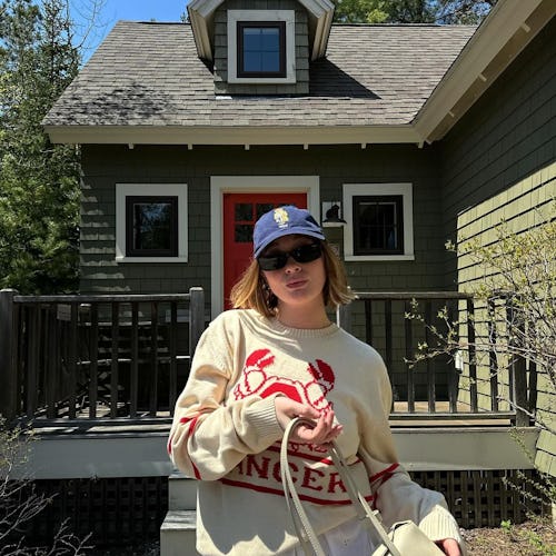 Woman in a cream sweater and cap stands in front of a green house with red door.