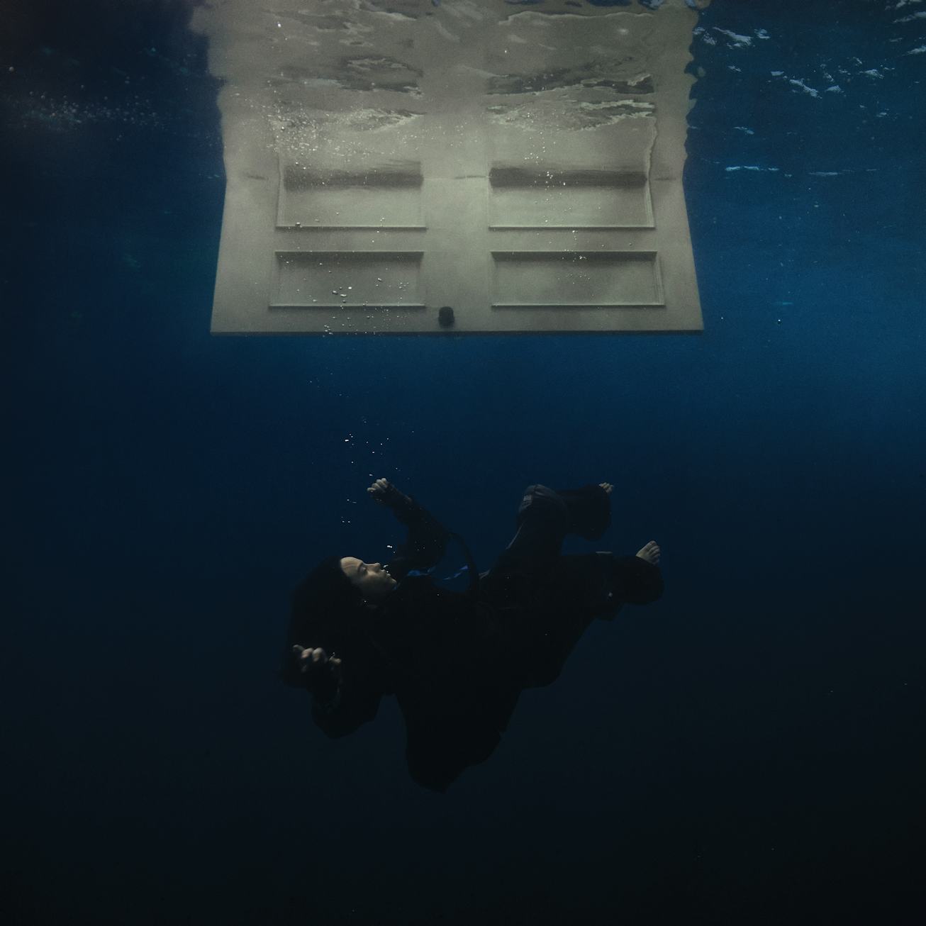 A person in a wet suit floats underwater facing a translucent floating object that reflects light.