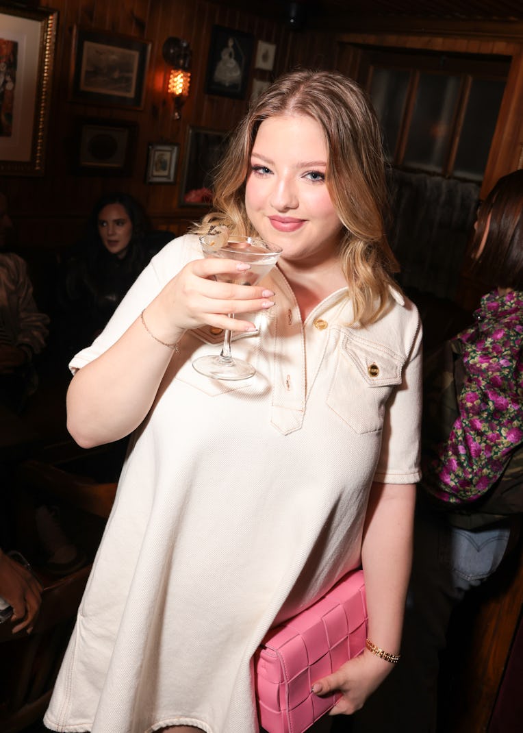 Young woman smiling at the camera, holding a cocktail, with a pink purse in a cozy bar setting.