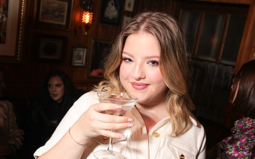 Young woman smiling at the camera, holding a cocktail, with a pink purse in a cozy bar setting.