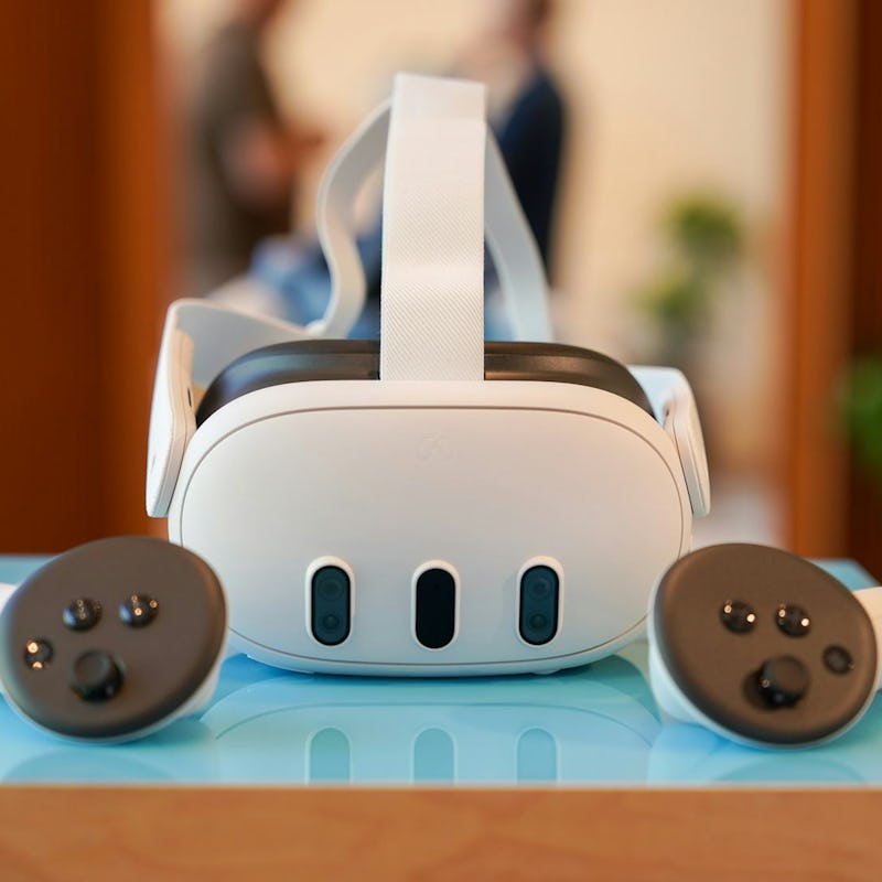A virtual reality headset and its two handheld controllers on a table, with a blurred figure in the ...