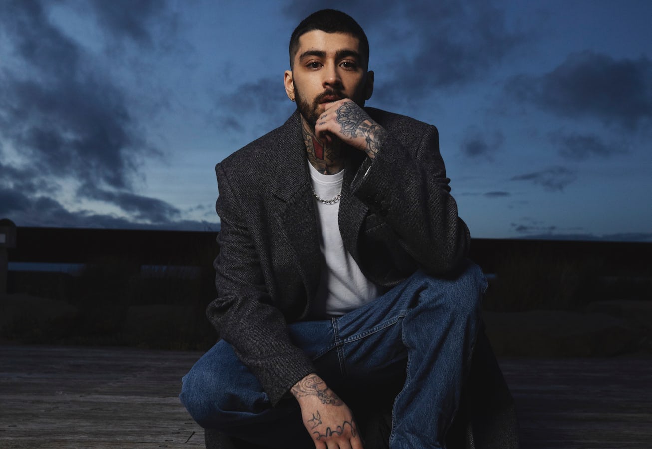 Man with tattoos crouching on a wooden platform, wearing a grey jacket and blue jeans, with a cloudy...