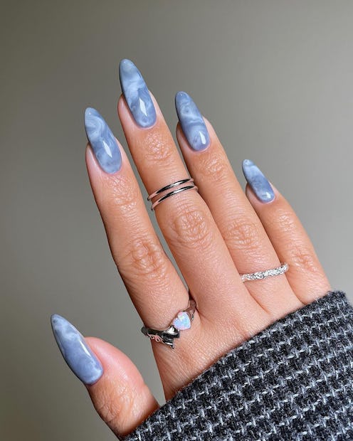 Close-up of a hand with long, almond-shaped, marble-effect blue nails and multiple silver rings, aga...