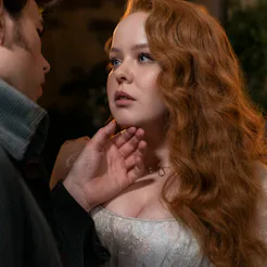 A woman with curly red hair, looking intently at a man who gently holds her chin, both illuminated s...